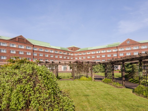 An exterior view of the Golden Jubilee Hotel shows a long 4 storeyed, red brick building. The image shows 2 arms of the hotel stretching the width of the image. In the foreground lawns, shrubs and trellises decorate the grounds. The building is pictured against a blue sky. 