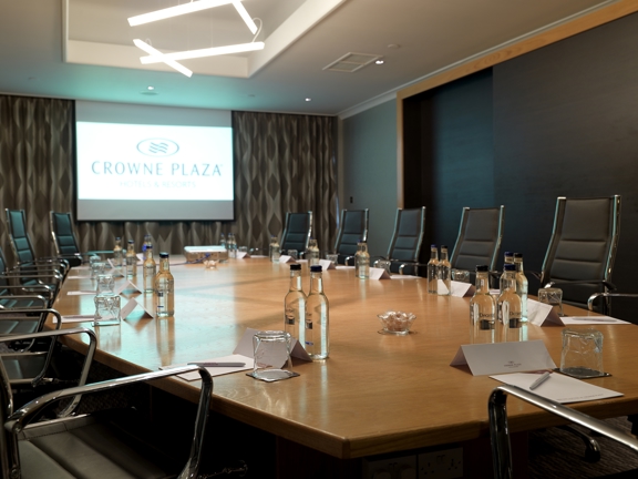 An interior view of Crowne Plaza's boardroom. The image is dominated by a large wooden table with 6 edges; around which are a dozen black and chrome high-backed chairs. The far wall is decorated with dark, patterned wallpaper and part of a modern light fitting can be seen on the ceiling. The room is otherwise lit by a projected screen, which is on the furthest wall; the projected image reads "Crowne Plaza". Glasses, name cards, paper and pens are all visible on the table.