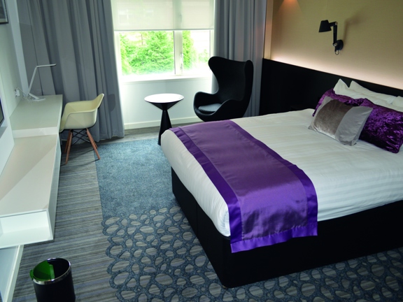 An interior view of a double room at the Golden Jubilee hotel. The room has a geometric carpet in muted colours, there is a double bed with crisp white sheets and a purple throw against the right wall. On the left is a narrow white desk, a wall-mounted television, a desk lamp and a white formed-plastic chair. Next to the curtained-window on the far side f the room is a stylish arm chair and a small coffee table. Wall lamps and framed prints are also visible.