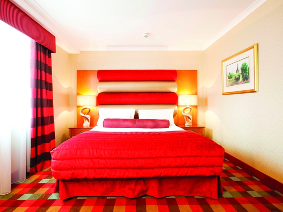 An internal view of a Carlton George bedroom. The room is decorated with a brightly coloured plaid carpet and a red headboard. The bed is decorated with a red cushion and blanket. There is a framed print on the right wall and a window with red curtains on the left.