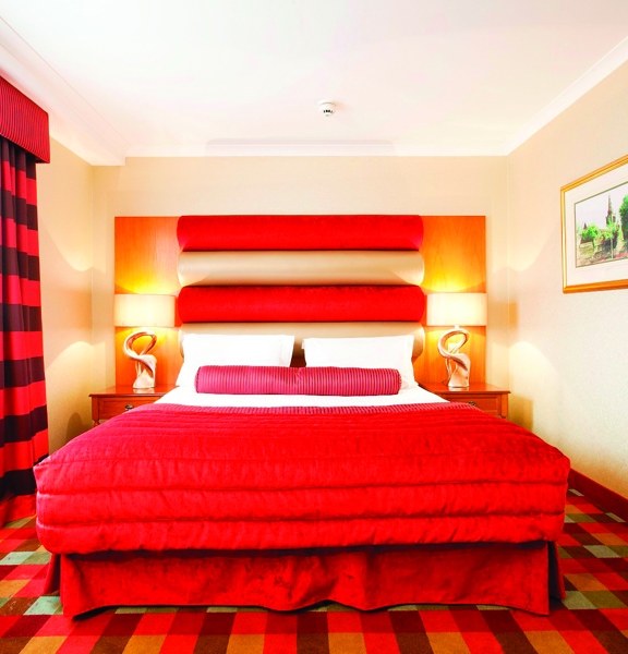 An internal view of a Carlton George bedroom. The room is decorated with a brightly coloured plaid carpet and a red headboard. The bed is decorated with a red cushion and blanket. There is a framed print on the right wall and a window with red curtains on the left.