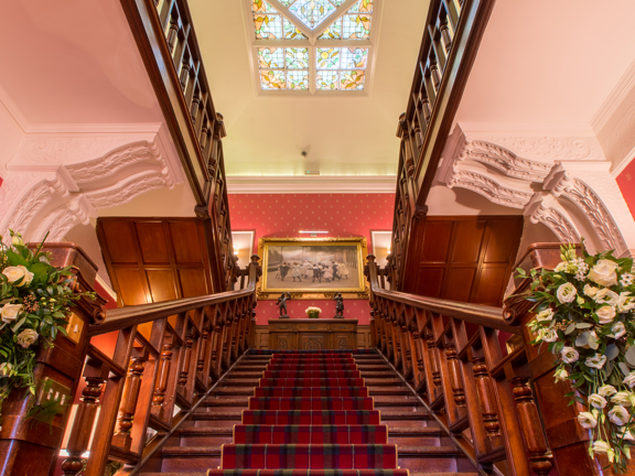 Wooden staircase with red tartan carpet, floral decorations on the bannister and large stained glass window above