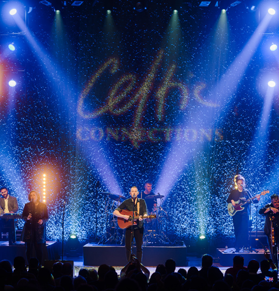 Folk band perform on stage in front of a blue textured background featuring the Celtic Connections logo.