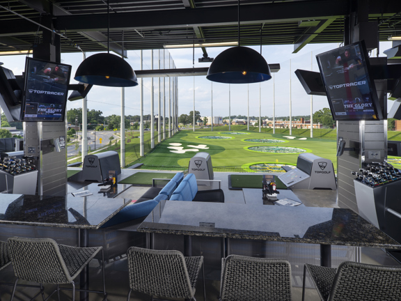 A photo looking out at the TopGolf driving range. A grey, metal awning stretches out over an area of seating with tables, screens and tees. The seating is a mix of low, blue sofas with coffee tables and grey, woven seats at marble counters- that look out to the course. The floor is grey and looks like concrete and the tees are astro turf. In the distance the greens, sand bunkers and net fences of the driving green can be seen. Buildings and roads can be seen near by, it is daytime and the sky is cloudy.