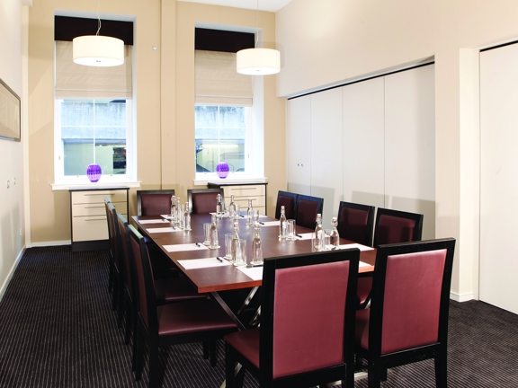 An interior view of the Fraser Suites meeting room, a bright room with 2 windows, with blinds, in the far wall. The room has white walls and a dark grey carpet. The room has a large rectangular wooden table and a dozen high-backed, wooden and leather-effect chairs around it. There are 2 purple glass vases on the windowsills and on the table paper, glasses and water bottles. On the wall there is a framed print and plug sockets visible.