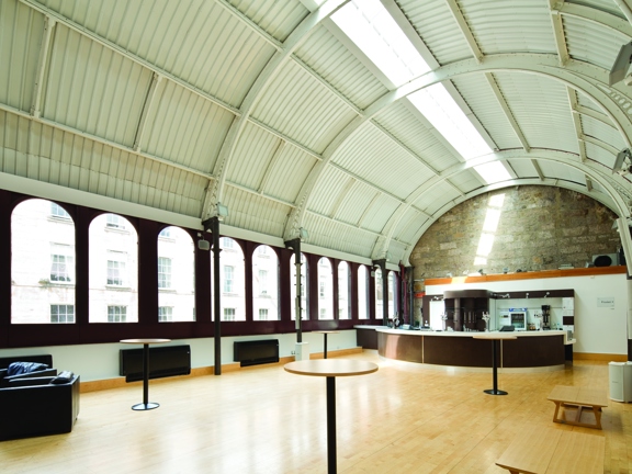 An interior of the Old Fruitmarket and City Halls, Candleriggs Bar. A space, brightly lit by daylight with a curved, vaulted ceiling and polished wood floors. Large arched window line the length of the left wall and a narrow skylight runs the length of the roof. At the furthest end of the room a brown and white semi-circular bar sits against the wall. The far wall's arched height is bare stone but otherwise the walls are mainly white. Leather arm chairs and high wooden tables are dotted around the room.
