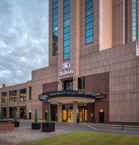 An exterior view of the Glasgow Hilton Hotel, shows a modern building with pinkish stone facing, there is a tarmac driveway, lawn and paved walkways also visible. Golden light can be seen through the glass main entrance; a large, lit, white Hilton logo  sits on the facade above a semi-circular glass awning. The ground & first storey stretch out to the left and right.  A broad, multi-storey tower of the same stone and glass rises out of the centre of the building & continues off the edge of the image.