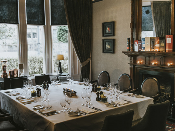 An internal view of a private dining space at Hotel du Vin shows a large square table laid for dinner surrounded by 10 wood and velvet chairs. The room feel opulent with a grand, wooden fireplace on the left-hand wall, a large mirror sits above the mantel which is lined with whisky boxes; small candles sit on a carved shelf below. The dark, fabric blinds on the bay window are half down but the heavy curtains are tied back. Small framed paintings decorate the subtly patterned wallpaper further.