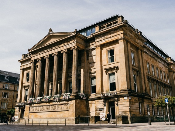An exterior view of Citation shows an imposing, golden-sandstone building adorned with carved details and 6 large, Greek Ionic style pillars. Windows are visible on 4 storeys and grey lettering reading "Citation" is visible above doors on either side of the pillars.