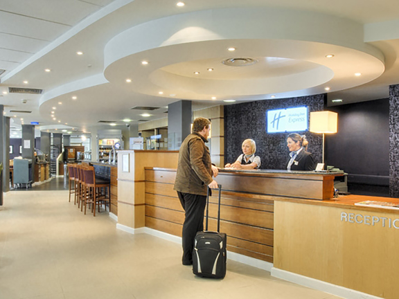 An interior view of the reception area at Holiday Inn Express, Riverside, shows a bright open-plan entrance way. On the right a light-wood tiered desk is attended by 2 smartly dressed women, a man with a suitcase is seen talking to them. Beyond the desk a long bar with high stools can be seen, pillars with tables and catering equipment in the background suggest a restaurant space beyond. The light suggests the left hand wall has large windows. Potted plants, a TV screen and lighting decorate the space., 