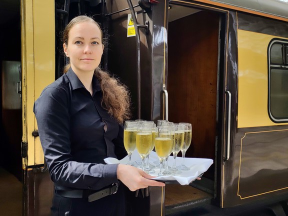 The image shows a young woman with dark, tied-back, hair stood at the entrance of an old steam train carriage. She is dressed in all black and holds a tray of champagne flutes. The train carriage is painted in brown and cream, and the interior wall visible is varnished wood. A small black metal step is visible between the door and the platform, which has a white edge and tactile tiling. An arching, glass roof is visible above.