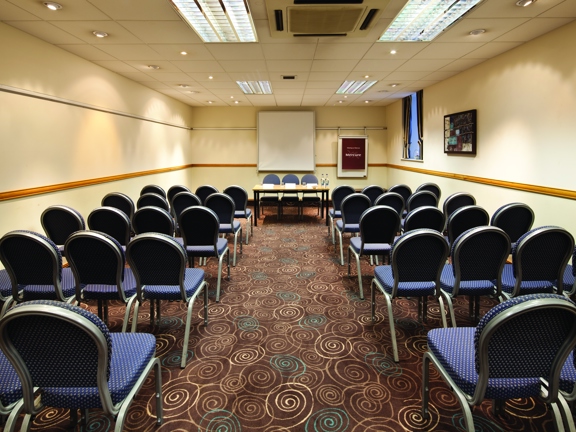 A Mercure hotel interior of the Broker Suite meeting room. The room is carpeted with a patterned brown carpet. The walls are cream, the ceiling is tiled and has fluorescent lights set into it. The room is filled with chairs arranged in rows. 5 rows can be seen with an aisle down the centre. At the far end of the room, where the chairs are facing, a whiteboard, flipchart, a table with 3 chairs are all visible. To their right is a curtained window. Framed prints can also be seen on the right-hand wall.