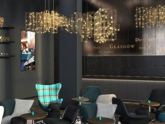 An interior shot of the Motel One Lounge depicts a room with dark grey walls and ashen, wooden floors. There is a variety of small tables and plush seats in velvet and wool textures and shades of teal and grey. A modern wing-backed chair in vibrant, turquoise tartan is most notable. The walls are decorated with shelves, gold letters read "Glasgow" on one and a golden locomotive is visible on another. The ceiling lights are a feature, consisting of cubes and grids of golden wire gleaming with little lights.