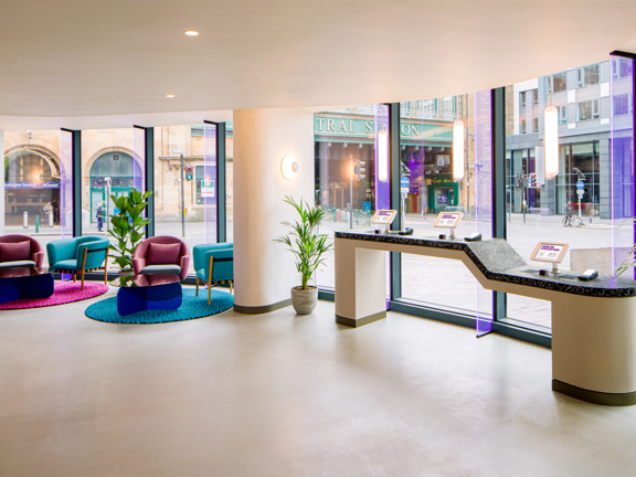 The image shows the bright, futuristic lobby of Yotel. A ground floor, curved, glass-sided space looking out at a street corner and the Glasgow Central Station bridge. The floor is pale and looks smooth, 2 rounded pillars and the low, spotlight-studded ceiling are rendered in bright white. 2 small seating areas of modern armchairs, round textured rugs and low glass coffee tables, in shades of purple and turquoise, are to the left. A narrow terrazzo counter with 3 mounted tablet screens is on the right. 