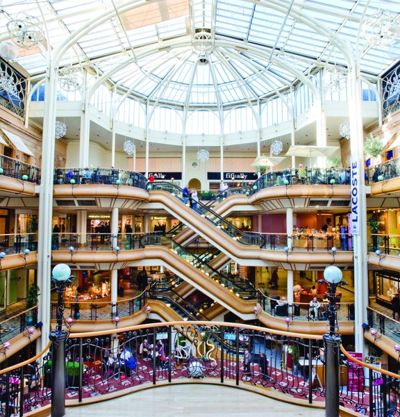 An interior image of the vast, tiered interior of Princes Square and its impressive pitched glass roof. Curving wooden balconies with black railing and sleek elevators, sweep around the outside edges of the open atrium. Awning, signs and banners advertise the shops, restaurants and businesses housed within Princes Square. It is a bright day and daylight spills through the roof. 