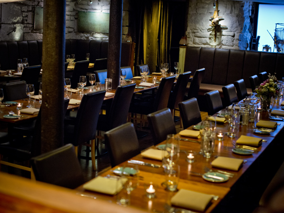 An image of Stravaigin's downstairs dining space. The space is filled with long, glossy, wooden dining tables, (laid for dinner,) the long edges are lined  with leather, high-backed chairs. The space has bare stone walls lit with spotlights. 