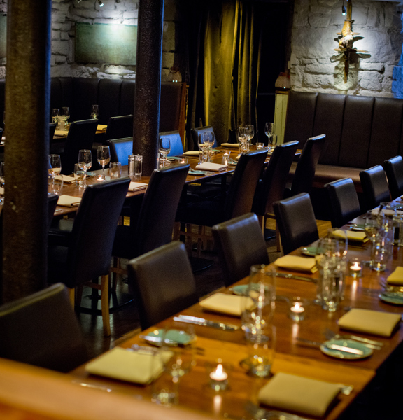 An image of Stravaigin's downstairs dining space. The space is filled with long, glossy, wooden dining tables, (laid for dinner,) the long edges are lined  with leather, high-backed chairs. The space has bare stone walls lit with spotlights. 