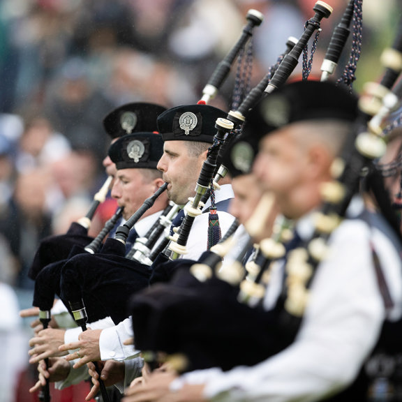 A group of bagpipers stand in a line in traditional clothing, playing in unison.