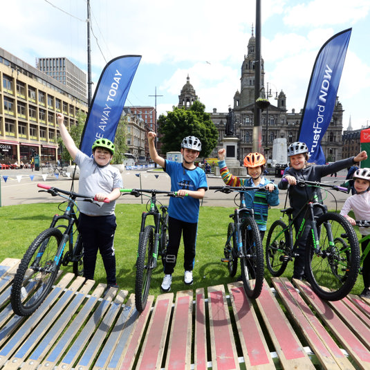 Five children stand next to their bikes cheering, in George Square, Glasgow.