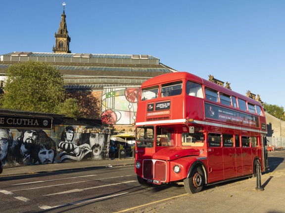 An image of the Red Bus Bistro, vintage double-decker bus parked beside Glasgow's famous Clutha Bar, its tree and its painted murals. It is a beautiful sunny day with blue skies. An old, stone spire and the grand, windowed roofline of the Briggait, a former fish market, re visible beyond. 