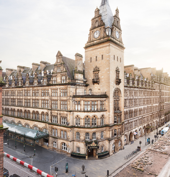 A view of the voco Grand Central buildings exterior from a high viewpoint over the road. A grand blonde sandstone hotel with chimney stacks, a large clock tower, gables and carved details; takes up the far corner of a road junction. The pavement on either side of the building is wide and paved, bike racks are visible. A long glass awning can be seen on the groundfloor of the buildings right hand facade. The red sandstone building the photo is taken from and the top floors of one across the road are visible.