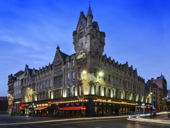 An exterior view of the Fraser Suites shows a Victorian sandstone building on a street corner, with a grand baronial inspired turret and carved details on the facade. The image is taken at dusk with a long exposure so streaks of light fill the road; streetlights and lights and signs from the ground floor businesses also light the scene. More modern buildings neighbour the Fraser Suite building at either end. 