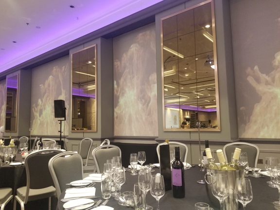 An interior view from a Hilton Hotel event space. Shows one edge of a room, the ceiling is uplit with a lilac glow and projections on the walls resemble flames. The wall has intermittent mirrored panels and are painted a pale grey. There are 2 round tables in view, covered with black table cloths and surrounded with pale-grey chairs. There are wine coolers and glasses placed on each table. A large speaker on a stand is visible on the right of the image, behind the second table.