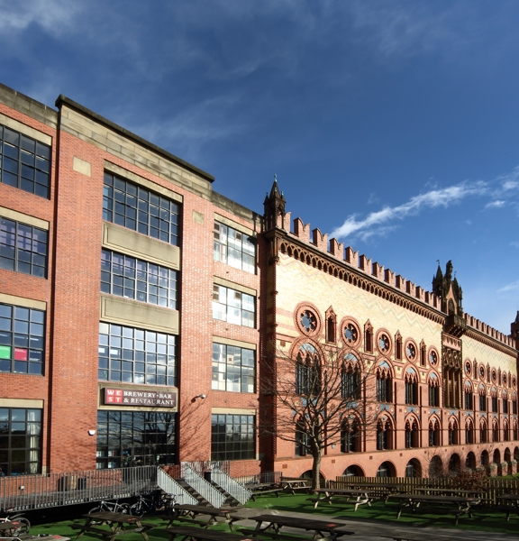A photo of the West on the Green brewery distinctive exterior. The oldest, original part of the building was inspired by Venetian palaces. The facade is made of red and yellow bricks, arranged into geometric patterns. A later red brick extension with large landscape, dark-framed windows take up large portions of the visible facade. A small set of metal stairs lead down to a grassy beer garden with picnic tables. A verdent corner of Glasgow Green is visible beyond the beer garden fence.