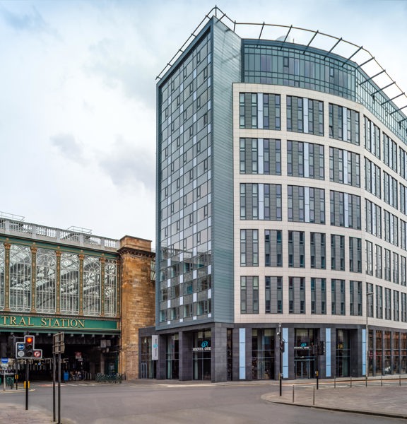 An exterior view of the Motel One building shows a modern, glass and cream, multi-storey building. The building is on the furthest corner of a cross roads, pedestrian crossings are visible over each of the 4 roads, bike racks and public bins are also visible. The hotel adjoins, Glasgow Central Station. A stone bridge with dark green signage and gold lettering goes over the farthest road of the cross-roads, perpendicular to the hotel entrance. The station has huge arched windows the length of the bridge.