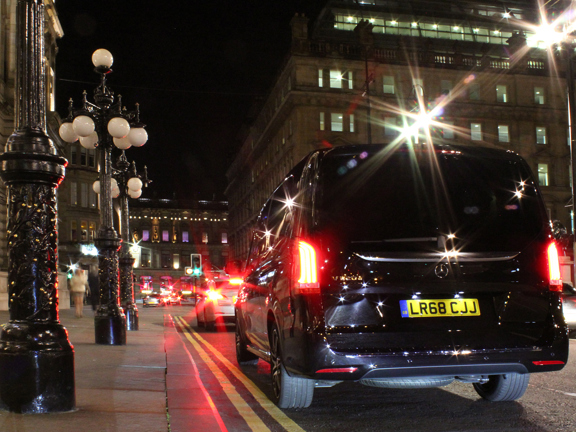 An image of the back of a boxy, black people carrier parked at George Square at night. Ornate lampposts line the payment on the left and in the distance the street is lined with large sandstone Victorian buildings. Car break lights and modern lampposts have created lens flares on the image.