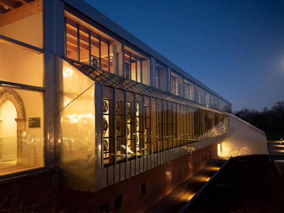 An external view of The Burrell Collection showing the long glass wall of the main museum lit up at night. A stone archway and stained glass windows are visible inside the museum, while a lit path and the moon are visible in the background.
