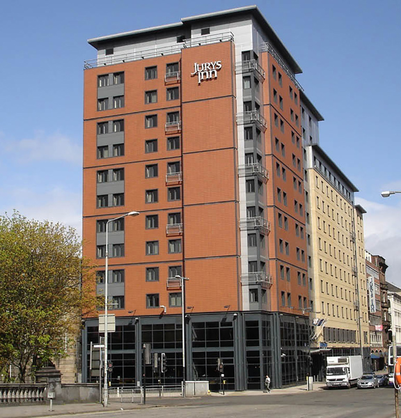 An external view of the Leonardo Hotel, shows a multi-storey, modern, red-brick building positioned on the corner of a junction. Metallic balconies punctuate the facade. The ground floor is windowed with dark glass and frames from floor to ceiling. A mixture of modern and Victorian buildings can be seen neighbouring the hotel on either side. A large, leafy tree and the carved stone of a bridge can be seen on the left. 