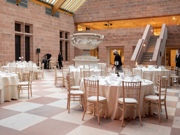 An internal view of the main foyer of The Burrell Collection set up for a dinner event. A large space with a checkerboard stone floor. Round tables with white table cloths surround the Warwick Vase, a large, marble urn. Speakers, lighting and technicians are also visible. A staircase to the first floor is visible in the background.