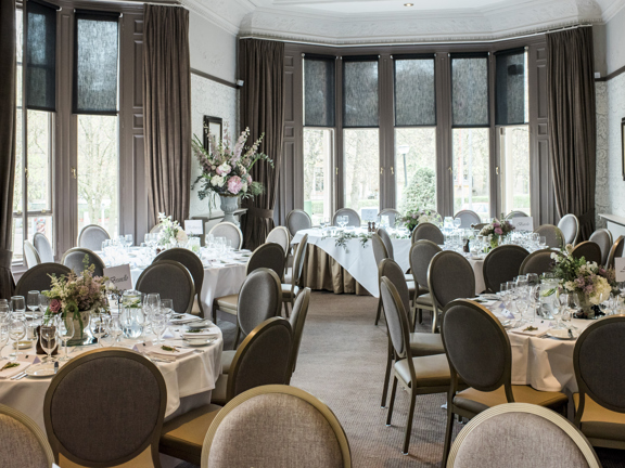 An interior view of Hotel Du Vin showing a room decorated for an event. Bays of wooden sash windows make the room, carpeted in neutral textured carpet and a subtle, floral wallpaper, bright and airy. A dark wood picture rail runs around the wall, matching the carved wooden and grey velvet chairs that surround round tables with crisp white table cloth, filled with flowers, glasses, silverware and china. A long, rectangular top table can be seen at the far end of the room. Framed prints decorate the walls.