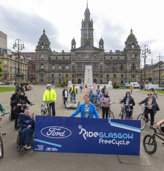 A group of cyclists pose with a blue Ride Glasgow banner in George Square, Glasgow.