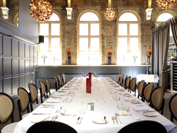 An interior view of a private dining space at Metropolitan, shows a bright room, the far wall is bare sandstone and has arched windows with cream frames. The left hand wall is dark brown and panelled. A door and speaker are visible at the far end of the wall. A large table, set for 20, is decorated with crisp white table cloths, silverware and crockery. Dark wood and velvet chairs with round backs surround it. On the right a banister suggests a step or two up to the main restaurant beyond a curtained wall..
