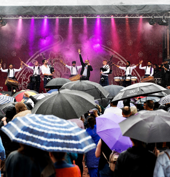 A crowd holding umbrellas watch Glasgow Mela performers on stage.