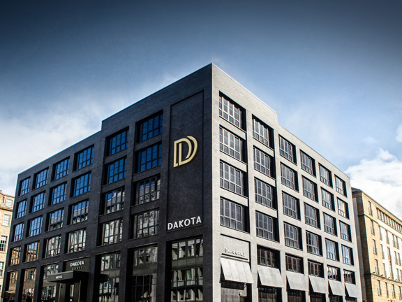 An exterior image of the Dakota Deluxe hotel shows a black, brick building with large black-framed windows on 6 storeys. A large gold logo depicting 2 overlapping Ds dominates the left hand side facet of the building, there is also smaller white lettering reading "Dakota". The building is framed against a blue sky and neighbouring it on either side are sandstone buildings.