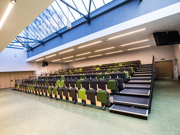 An interior view of one of Glasgow Caledonian's conference halls. The image shows a brightly lit room with theatre style seating on a stepped platform. The seats are in varying shades of green and grey. The roof is vaulted with grey metal beams visible against a large skylight. Ceiling lights are also on and a large television screen and speakers can be seen suspended from the ceiling. In the bottom right corner the double door entrance can be seen.