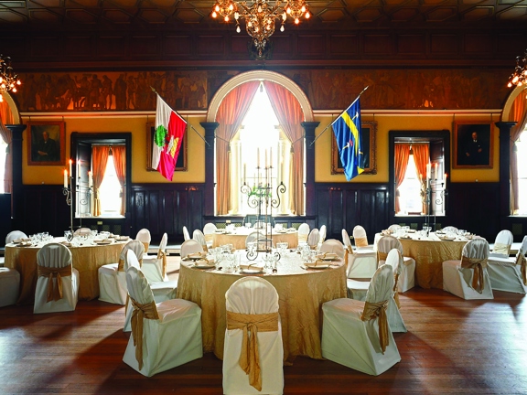 A photograph of the Trades Hall, Grand Hall decorated for a private dinner. The room has a dark wooden floor and panelled walls. The ceiling is also wooden, with gilt edges defining the hexagonal carving. A guilt border of Grecian-style figures runs between the ceiling and picture rail. Light pours through an arched window, (adorned with 2 flags,) and 2 smaller rectangular windows. Round tables and chairs are decorated with gold cloths & details. 