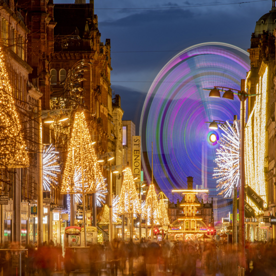 Shopping street lit up for the festive season, with shoppers blurred. Illuminations include cone shaped structures. Large lit up ferris wheel blurred in the background