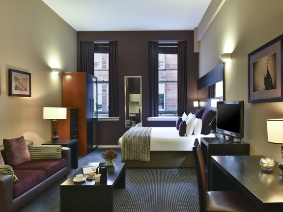 An interior view of a Fraser Suites studio. The far wall is painted a deep purple and has 2 windows - with dark pulled-back curtains; there is a long mirror between them. The other walls are cream and have framed prints on them. In the far-right is a large, clean double bed. Closer, on the left, there is a deep purple sofa & a coffee table. On the right there is a desk; the furniture is black or dark wood. A large television sits on a stand between the desk and the bed, lamps and sockets dot the room.