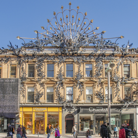 People strolling along shop-lined Buchannan Street, in front of Princes Square shopping centre which is covered in large wrought iron foliage and has a 10m wide stylised iron peacock on top.
