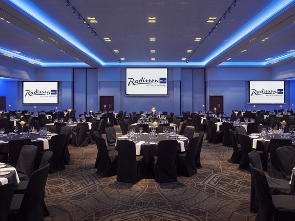 An interior image of the Megalithic Suite at Radisson Blu shows a large event space with a grey mandala patterned carpet. Pale blue lights light the ceiling and walls. 3 large projects or screens displaying the Radisson Blue logo also light the room.  About 20 large round tables are surrounded by chairs. They are covered with black cloths, wine glasses, cloth napkins and candles decorate the tables. 3 double doors are visible in the far wall.