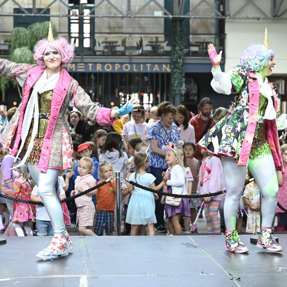 Two performers wearing unicorn horns and colourful costumes dance on stage in front of a crowd of children in Merchant Square.