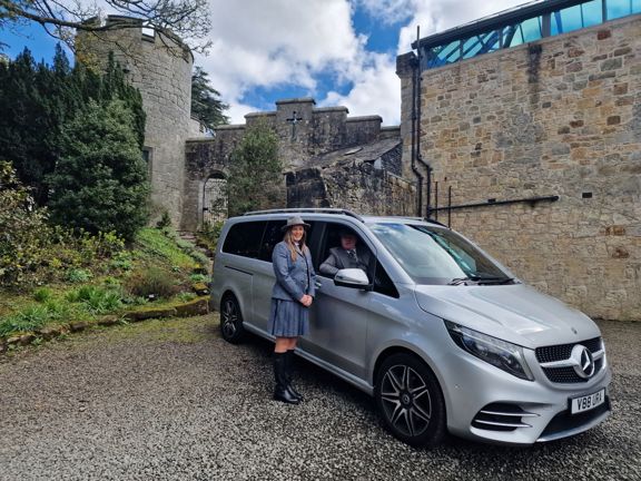 Woman standing next to a silver Mercedes people carrier wearing 3 piece tweed skirt suit. Man seated in driving seat with window open. Ancient sandstone building behind.