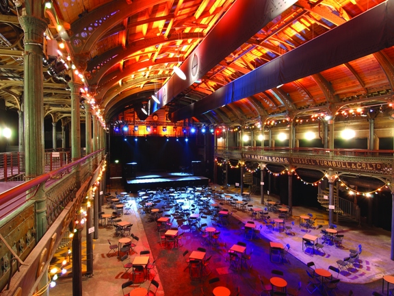 An interior view from Old Fruitmarket & City Halls shows a large, 2 storey hall with a vaulted ceiling. The photo is taken from a first storey balcony, ornate iron pillars, arches and railings are visible around the hall, balcony and ceiling. The structure of the ceiling is lit with golden lights; spotlights and festoon lights around the perimeter also light the space. Bistro tables and folding chairs are arranged in small groups in front of a large, black stage with a technical lighting rig.