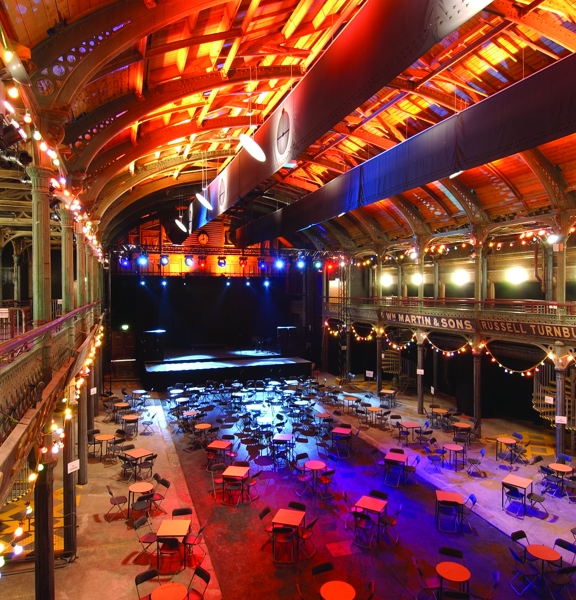 An interior view from Old Fruitmarket & City Halls shows a large, 2 storey hall with a vaulted ceiling. The photo is taken from a first storey balcony, ornate iron pillars, arches and railings are visible around the hall, balcony and ceiling. The structure of the ceiling is lit with golden lights; spotlights and festoon lights around the perimeter also light the space. Bistro tables and folding chairs are arranged in small groups in front of a large, black stage with a technical lighting rig.