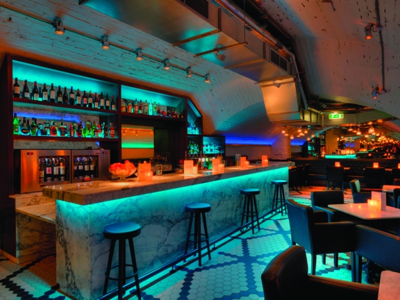 An image of the Alston's white, marble bar. The floor is tiled in small black and white tiles, laid in geometric shapes. The bar and the white, brick walls are up-lit with turquoise lights. 4 minimal black stools sit along the length of the bar, while small, low marble tables are surrounded by modern, brown leather armchairs to the right of it. Shelves line the curving wall behind the bar, they are also lit turquoise and are well stocked with bottles and glasses.