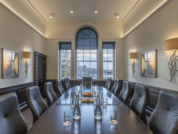 An interior view of  boardroom space at the Clayton Hotel. A long, dark table surrounded by plush grey chairs dominate the space. Glasses and bottles of water decorate the table. The walls of the room are decorated with modern light fittings and framed prints, while the end wall has a large, arched sash-window. Grey blinds, overhead lighting and a in the back left corner are also visible. 